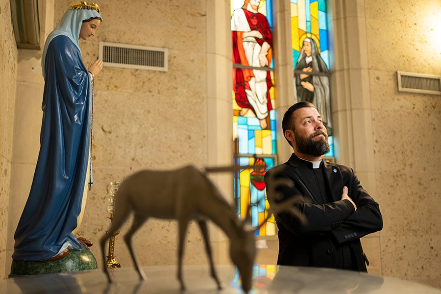 a priest looks into the light before a wooden deer, statue of Mary, a stained glass, and a cross.